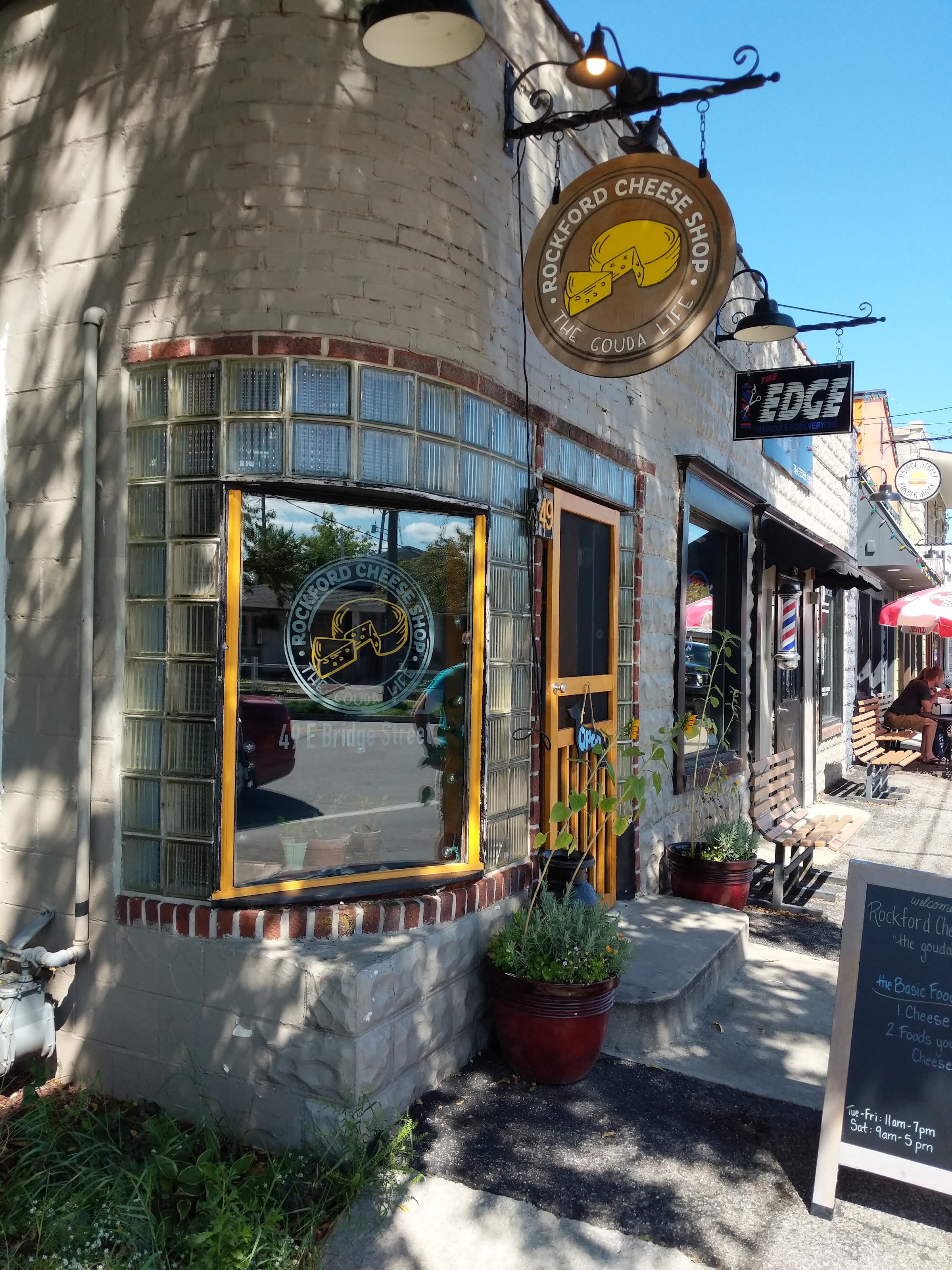 The Rockford Cheese Shop corner storefront on Bridge Street with its hanging round sign, glass-block windows, and a sandwich-board sign out front.