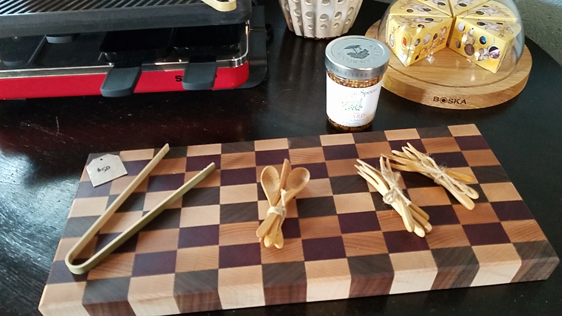 Cheese tools and a checkered serving board on a table — a raclette set in the background.