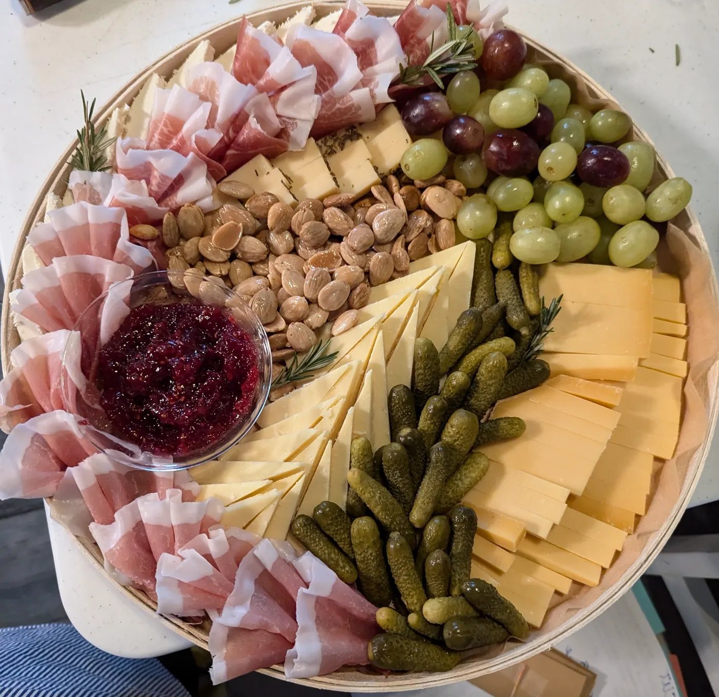 A round wooden board piled with sliced cheeses, prosciutto and cured meats, grapes, almonds, cornichons, and berry jam.