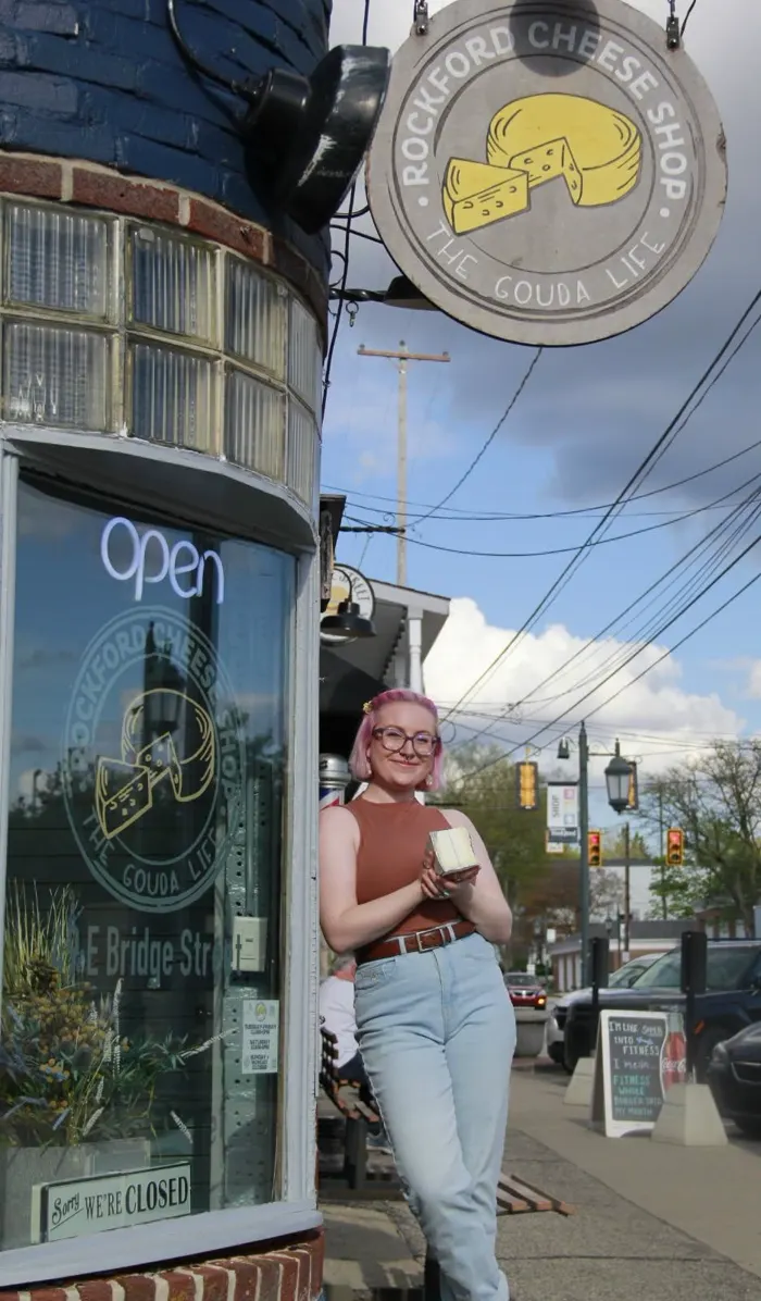 The Rockford Cheese Shop storefront on Bridge Street with its round hanging sign and a staffer holding a wedge of cheese.